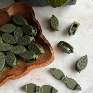 May include: A collection of green leaf-shaped clips in a wooden bowl. The clips are made of plastic and have a textured surface. There are also several other clips on a white surface, including some that are open and some that are closed.
