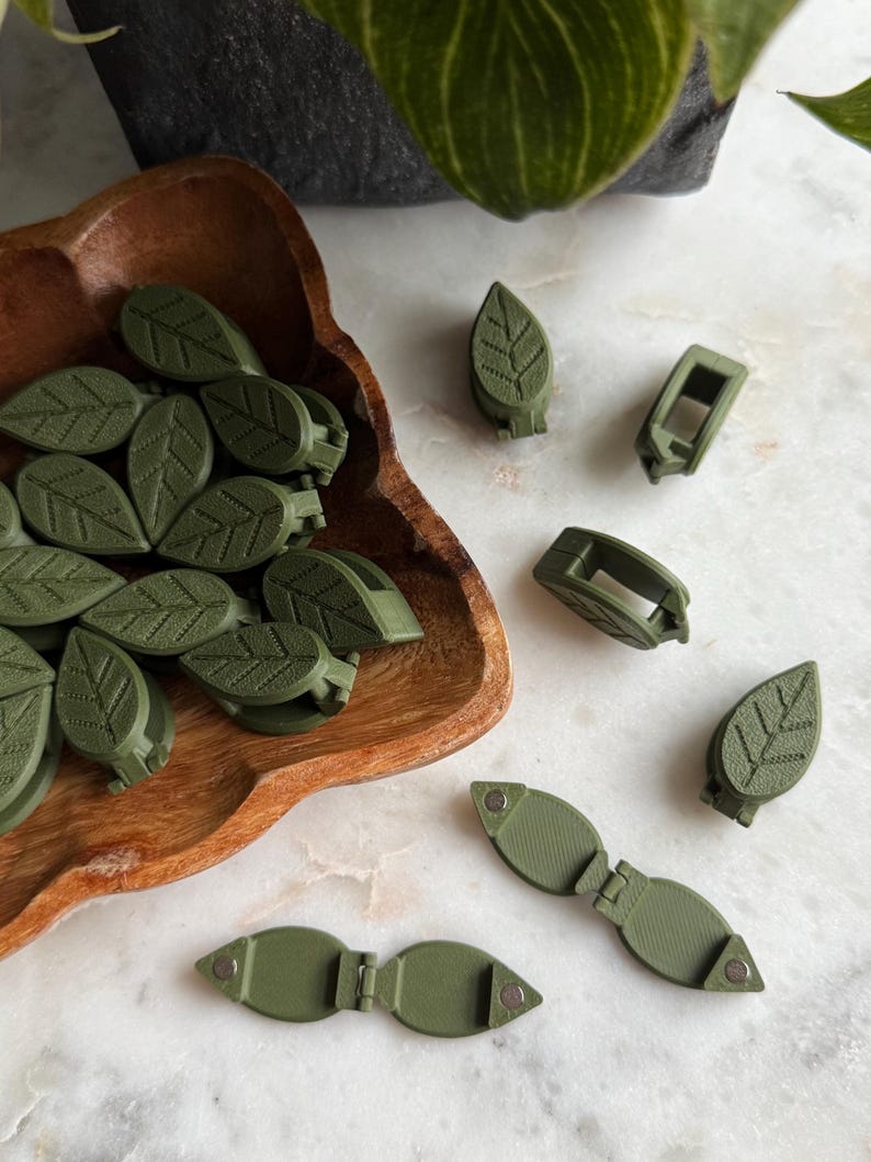May include: A collection of green leaf-shaped clips in a wooden bowl. The clips are made of plastic and have a textured surface. There are also several other clips on a white surface, including some that are open and some that are closed.