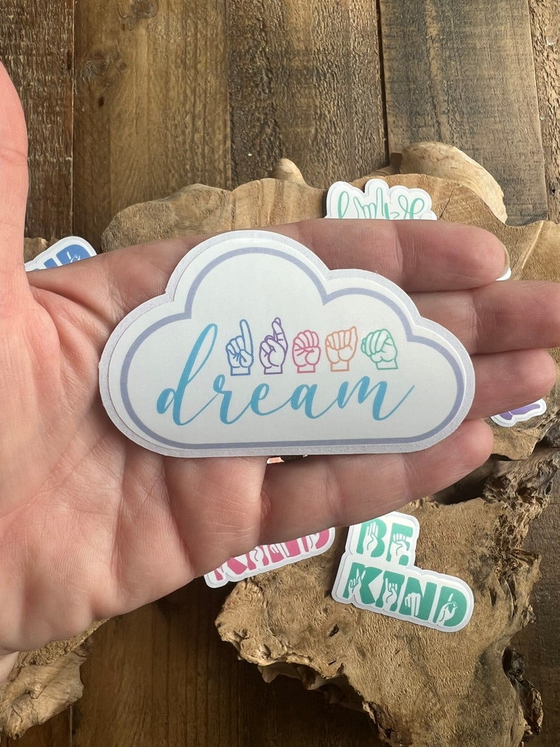Puede incluir: Una pegatina blanca en forma de nube con la palabra "dream" escrita en cursiva y cinco manos de colores arco&iacute;ris firmando la palabra "dream" en Lengua de Se&ntilde;as Espa&ntilde;ola.