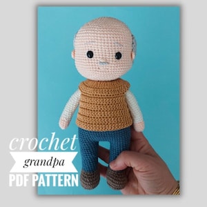 以下が含まれることがあります： おじいちゃんの編みぐるみパターン。茶色の髪、茶色のベスト、青いズボン、茶色の靴を履いたおじいちゃんの編みぐるみ人形。人形は手で持たれています。