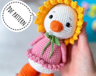 May include: A crocheted amigurumi duck wearing a pink dress and a yellow flower crown. The duck has orange feet and a green bow. The text "PDF PATTERN" is visible in the image.