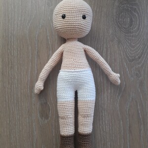 May include: A crocheted doll with a beige body, white shorts, and brown boots. The doll has black eyes and a simple face.