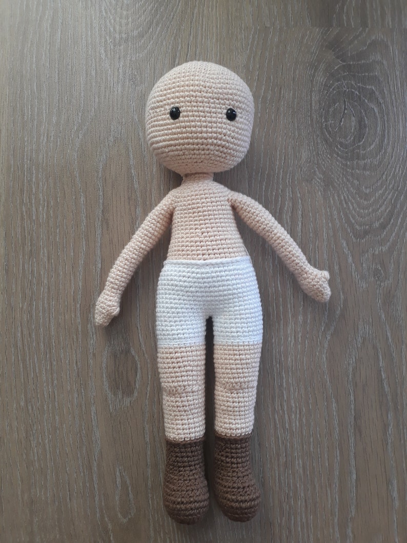 May include: A crocheted doll with a beige body, white shorts, and brown boots. The doll has black eyes and a simple face.