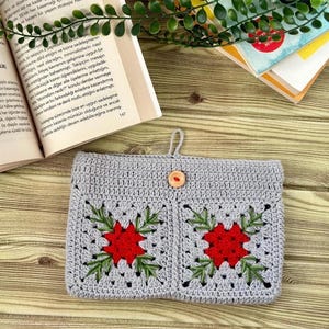 K&ouml;nnte beinhalten: Geh&auml;kelte graue Tasche mit zwei quadratischen Taschen, jede mit einer roten Blume mit gr&uuml;nen Bl&auml;ttern. Die Tasche hat einen Holzknopfverschluss und eine Schlaufe zum Aufh&auml;ngen. B&uuml;cher und Gr&uuml;nzeug im Hintergrund.