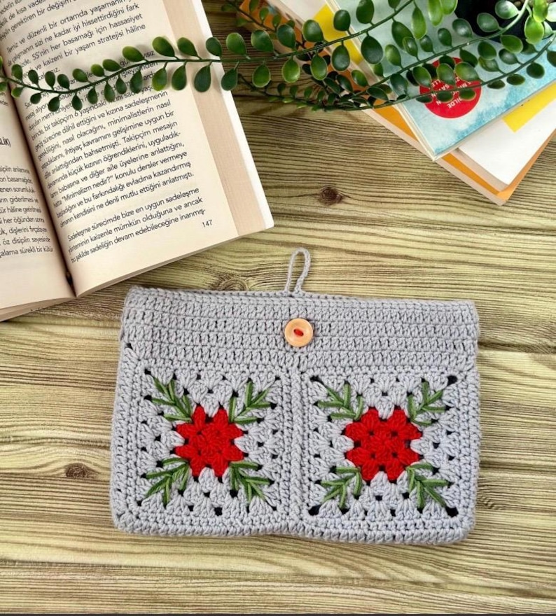 K&ouml;nnte beinhalten: Geh&auml;kelte graue Tasche mit zwei quadratischen Taschen, jede mit einer roten Blume mit gr&uuml;nen Bl&auml;ttern. Die Tasche hat einen Holzknopfverschluss und eine Schlaufe zum Aufh&auml;ngen. B&uuml;cher und Gr&uuml;nzeug im Hintergrund.