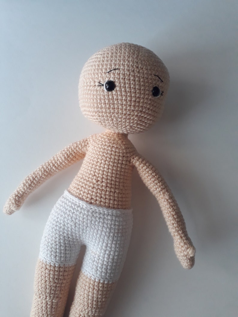 May include: A crocheted doll with a beige body and white shorts. The doll has black eyes and a simple embroidered face.