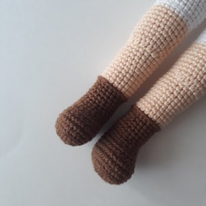 May include: Close-up of a pair of brown crocheted doll shoes. The shoes are made with a brown yarn and have a rounded toe.