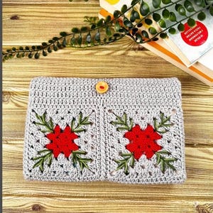 K&ouml;nnte beinhalten: Geh&auml;kelte graue Clutch mit zwei roten Blumenmustern und gr&uuml;nen Bl&auml;ttern. Die Clutch wird mit einem Holzknopf verschlossen. Die handgefertigte Tasche liegt auf einer Holzoberfl&auml;che, mit B&uuml;chern und Gr&uuml;nzeug im Hintergrund.