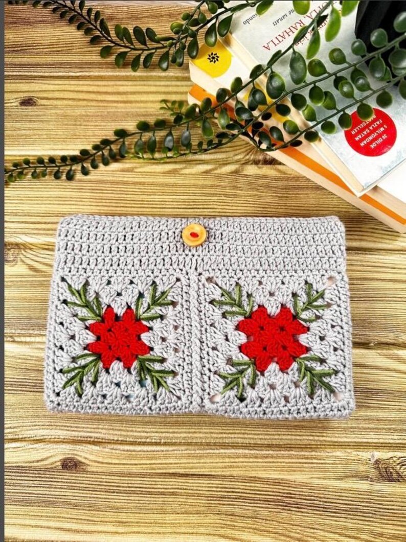 K&ouml;nnte beinhalten: Geh&auml;kelte graue Clutch mit zwei roten Blumenmustern und gr&uuml;nen Bl&auml;ttern. Die Clutch wird mit einem Holzknopf verschlossen. Die handgefertigte Tasche liegt auf einer Holzoberfl&auml;che, mit B&uuml;chern und Gr&uuml;nzeug im Hintergrund.
