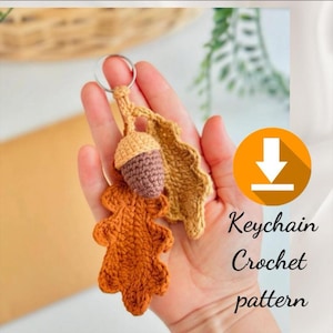 Può includere: Portachiavi all'uncinetto fatto a mano con una ghianda e due foglie nei toni del marrone e beige. Il portachiavi è attaccato a un anello d'argento. L'immagine include anche il testo "Keychain Crochet pattern".