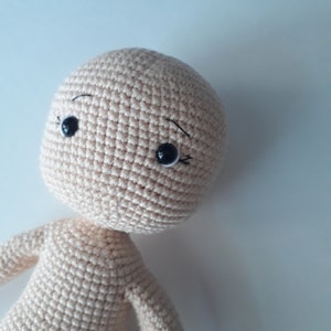 May include: A crocheted doll with a beige body and black eyes. The doll has a simple, sweet expression.