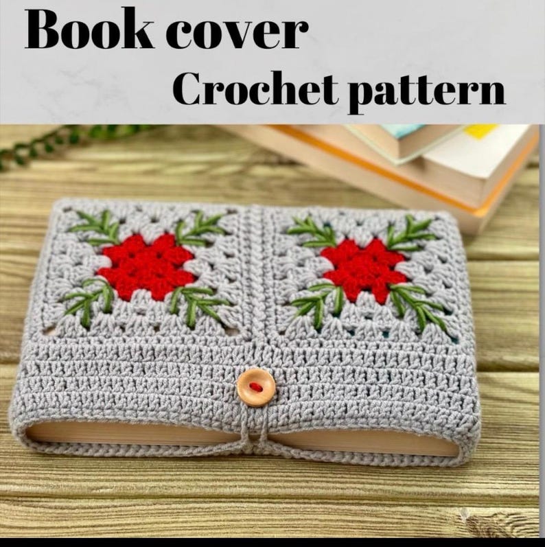 K&ouml;nnte beinhalten: Eine geh&auml;kelte Buchh&uuml;lle in Hellgrau mit einem roten Blumen- und gr&uuml;nen Blattdesign. Sie hat einen Knopfverschluss und den Text "Book cover Crochet pattern". Die Buchh&uuml;lle liegt auf einer Holzoberfl&auml;che.