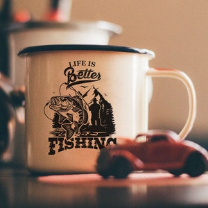 K&ouml;nnte beinhalten: Wei&szlig;e Emaille-Tasse mit einer schwarzen Grafik eines Fisches und dem Text "Life is Better Fishing".