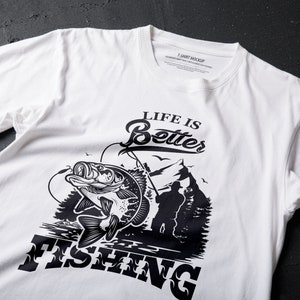 K&ouml;nnte beinhalten: Wei&szlig;es T-Shirt mit einem schwarzen Grafikdesign eines Fisches, der aus dem Wasser springt, mit dem Text "Life is Better Fishing".