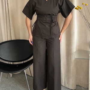 May include: A dark brown two-piece outfit. The top is a wrap style with short sleeves and a waist tie. The trousers are wide-legged. The outfit is made of a smooth fabric. The model is wearing light green shoes.