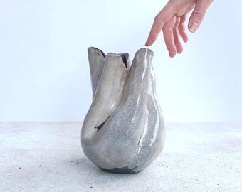 May include: A unique, handcrafted vase with a textured, metallic finish. The vase has an organic, flowing shape with a wide opening and a dark interior. The exterior is a blend of silver and gray tones, creating a modern, artistic aesthetic.