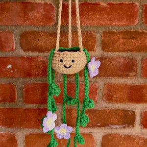 May include: A crocheted hanging plant decoration featuring a tan pot with a smiling face, green vines, and light purple flowers with yellow centers. The entire piece is suspended by three tan crocheted cords that meet at the top for hanging.