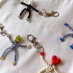 May include: A collection of beaded keychains in the shape of stick figures. The keychains feature various colors, including black, blue, white, and red. Some have star-shaped or heart-shaped accents. Each keychain has a silver-colored clasp.