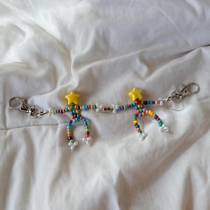 May include: Two beaded keychains in the shape of stick figures. Each figure has a yellow star head, rainbow body, and white feet. The keychains are connected by a magnetic clasp and have silver clasps for attaching to keys or bags.
