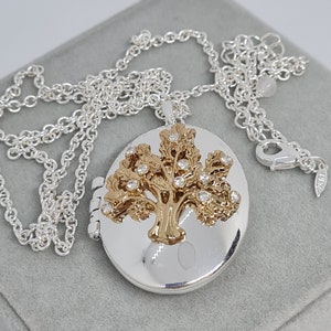 May include: Silver oval locket necklace featuring a gold-tone tree design adorned with clear crystals. The locket is suspended from a fine silver chain. The necklace is presented on a grey surface.