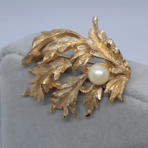 May include: A gold-toned brooch in the shape of stylized leaves, with a central cream-colored pearl. The leaves have detailed veining, and the brooch has a pin closure on the back. The design is reminiscent of nature-inspired jewelry.