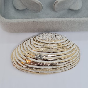 May include: A decorative seashell with a white and gold metallic finish. The shell has a ridged texture with alternating bands of white and gold. The top of the shell has a textured, speckled design. The shell is displayed on a white surface.