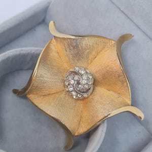 May include: A gold-toned metal brooch shaped like a five-petaled flower with a brushed texture on the petals. The center of the flower features a circular cluster of small, clear, sparkling rhinestones set in a silver-toned metal base.
