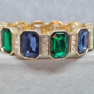 Könnte beinhalten: Ein goldfarbenes Gliederarmband mit abwechselnd smaragdgrünen und tiefblauen, rechteckig facettierten Steinen. Jeder große Stein wird von vertikalen Reihen kleiner, klarer, runder Kristalle flankiert, die in goldfarbene Metallfassungen eingefasst sind.