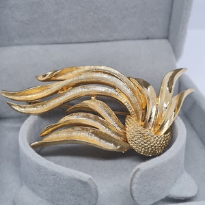 May include: A gold-toned brooch with a floral design. The brooch features layered, curved leaf-like elements radiating from a textured, spherical center. The brooch is set against a gray velvet background.