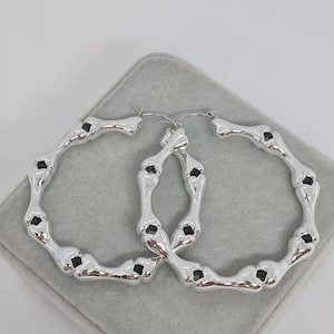 Puede incluir: Un par de pendientes de aro de plata con un diseño en forma de hueso. Los pendientes son grandes y tienen un acabado pulido.