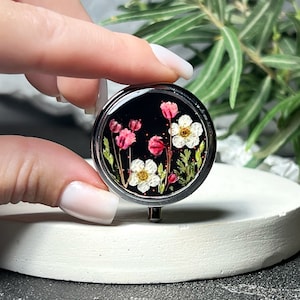 May include: A silver round trinket box with a black background and a pressed flower design featuring white and pink flowers.