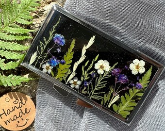 Peut inclure: Une petite boîte rectangulaire argentée avec un fond noir et des fleurs pressées à l'intérieur. Les fleurs sont blanches, bleues et vertes. La boîte porte une étiquette qui dit "Fait main" avec un cœur et un smiley.