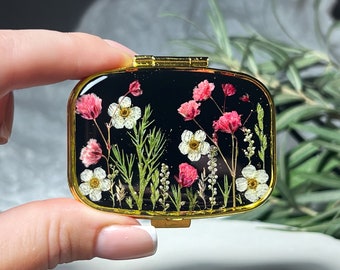 Op de afbeelding: Een klein, rond, goudkleurig doosje met een zwarte achtergrond en een ontwerp van geperste bloemen. De bloemen zijn wit en roze met groene bladeren.