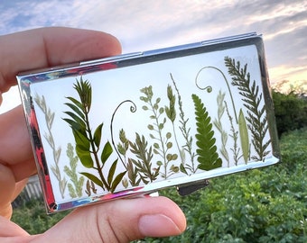 Peut inclure: Un porte-cartes argenté avec une fenêtre transparente affichant des plantes vertes et blanches pressées. Les plantes sont disposées verticalement en rangée.