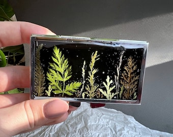 Peut inclure: Un étui en métal argenté avec un dessus transparent contenant des plantes vertes et brunes pressées sur un fond noir.