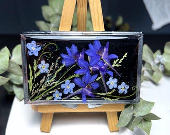 Peut inclure: Un compact en métal argenté avec un fond noir et un motif de fleurs pressées comprenant des fleurs bleues et violettes. Les fleurs sont disposées en grappe et semblent être pressées contre un fond noir.