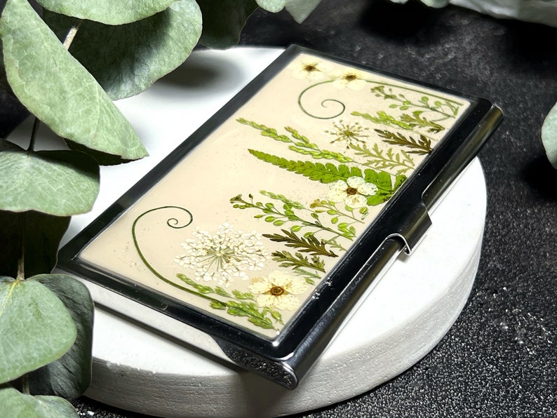 Op de afbeelding: Een zilveren visitekaartjeshouder met een cr&egrave;mekleurige achtergrond met geperste witte bloemen en groene bladeren.