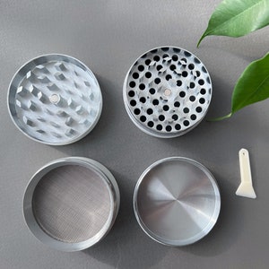 Pode incluir: Um moedor de ervas de metal prateado desmontado com quatro componentes separados. A pe&ccedil;a superior tem dentes em forma de diamante, a segunda tem v&aacute;rios orif&iacute;cios, a terceira uma tela de malha e a inferior &eacute; s&oacute;lida. Uma pequena esp&aacute;tula de pl&aacute;stico branco tamb&eacute;m est&aacute; inclu&iacute;da.