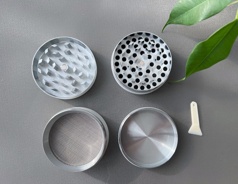 Pode incluir: Um moedor de ervas de metal prateado desmontado com quatro componentes separados. A pe&ccedil;a superior tem dentes em forma de diamante, a segunda tem v&aacute;rios orif&iacute;cios, a terceira uma tela de malha e a inferior &eacute; s&oacute;lida. Uma pequena esp&aacute;tula de pl&aacute;stico branco tamb&eacute;m est&aacute; inclu&iacute;da.