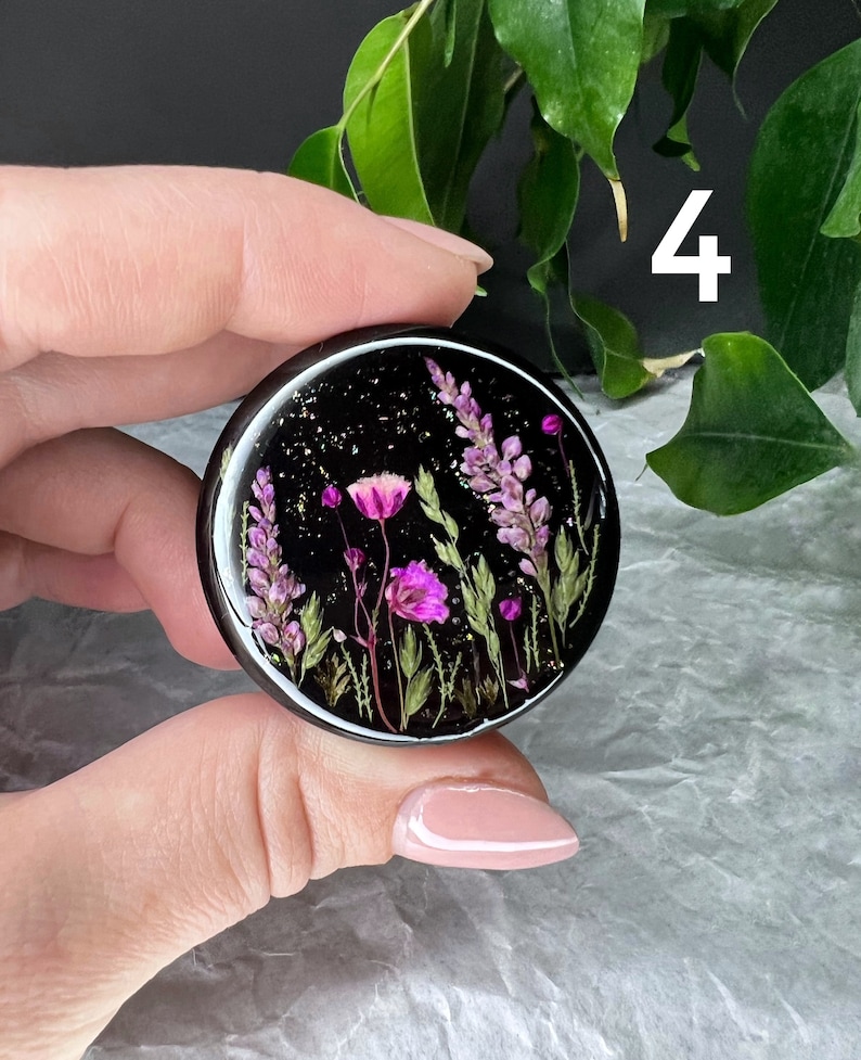 Pode incluir: Um objeto redondo de resina preta com um design floral. O design apresenta flores roxas e rosas, folhas verdes e flocos dourados. O n&uacute;mero "4" &eacute; vis&iacute;vel em branco no fundo.