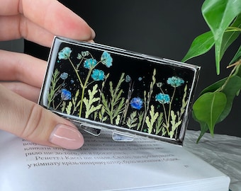 Peut inclure: Une boîte rectangulaire noire avec un dessus transparent remplie de fleurs bleues et vertes pressées et de paillettes. La boîte est posée sur un livre.