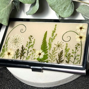 Op de afbeelding: Een zilveren visitekaartjeshouder met een beige achtergrond en geperste witte en groene bloemen en bladeren. De bloemen zijn in een verticale rij gerangschikt met een paar krulletjes boven en onder.