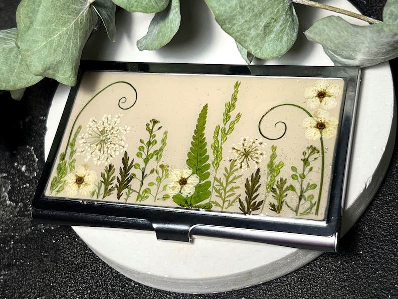 Op de afbeelding: Een zilveren visitekaartjeshouder met een beige achtergrond en geperste witte en groene bloemen en bladeren. De bloemen zijn in een verticale rij gerangschikt met een paar krulletjes boven en onder.