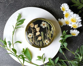 Op de afbeelding: Een ronde sieradenbox met een gouden rand, met een zwarte achtergrond en witte geperste bloemen erin. De doos staat op een wit bord, omringd door groene bladeren.