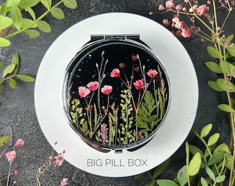 Op de afbeelding: Een ronde, zilveren compact spiegel met een zwarte achtergrond en een ontwerp van geperste bloemen. De bloemen zijn roze en groen en de tekst "BIG PILL BOX" is onderaan de spiegel gedrukt.