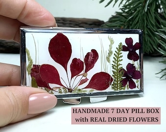 Peut inclure: Une boîte à pilules en argent avec un fond blanc et de vraies fleurs séchées pressées à l'intérieur. Les fleurs sont rouges, vertes et violettes. Le texte sur la boîte indique "HANDMADE 7 DAY PILL BOX with REAL DRIED FLOWERS".