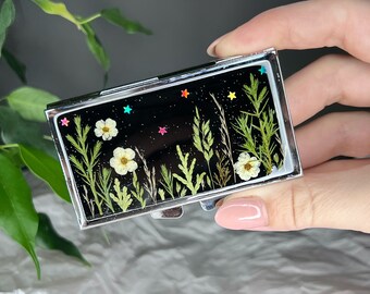 Peut inclure: Une boîte à pilules en argent avec un fond noir et un motif de fleurs pressées. Les fleurs sont blanches et vertes, et des étoiles irisées sont éparpillées sur le fond.