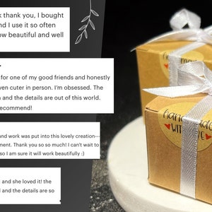 K&ouml;nnte beinhalten: Zwei kleine, quadratische Geschenkboxen aus braunem Karton, gebunden mit wei&szlig;em Band und einem Aufkleber "Handgefertigt mit Liebe". Die Boxen stehen auf einer wei&szlig;en Oberfl&auml;che mit gr&uuml;nen Bl&auml;ttern im Hintergrund.