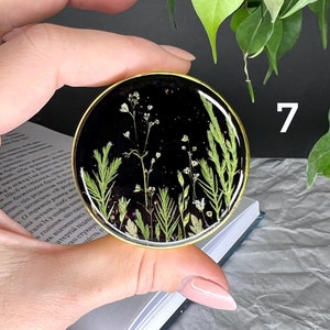 Pode incluir: Uma base de resina preta redonda com uma borda dourada, com flores secas e vegeta&ccedil;&atilde;o. A base &eacute; segurada em uma m&atilde;o. O n&uacute;mero 7 &eacute; vis&iacute;vel na imagem.
