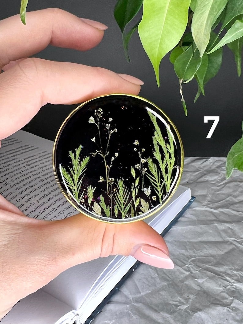 Pode incluir: Uma base de resina preta redonda com uma borda dourada, com flores secas e vegeta&ccedil;&atilde;o. A base &eacute; segurada em uma m&atilde;o. O n&uacute;mero 7 &eacute; vis&iacute;vel na imagem.